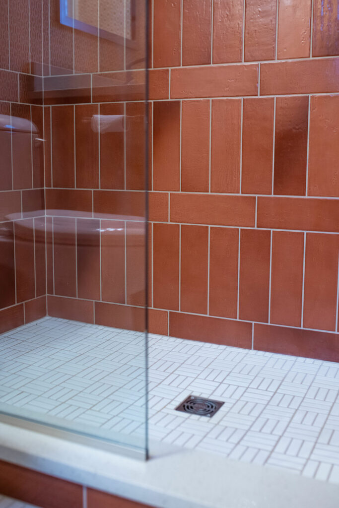 White shower floor tile, glass shower door, and orange wall tile in bathroom design. Lindsey Putzier Design Studio Hudson, OH