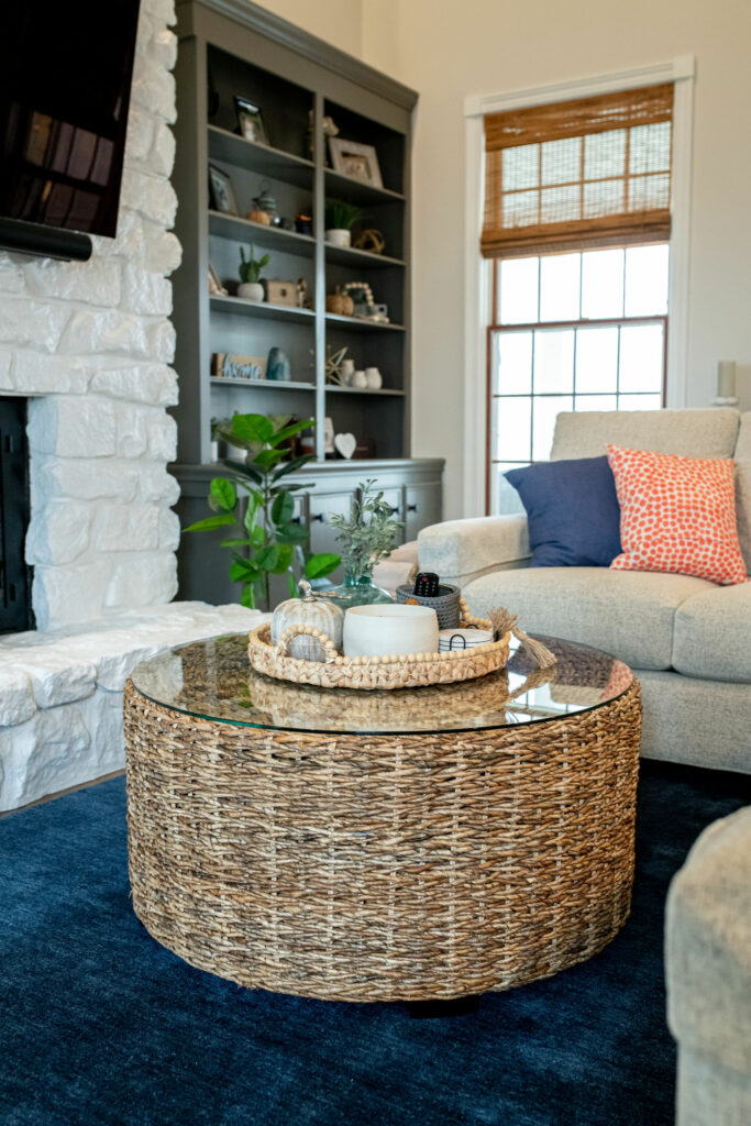 White bricked fireplace, natural shades, dark built-in shelves, and woven rattan ottoman in Family Room design. Lindsey Putzier Design Studio