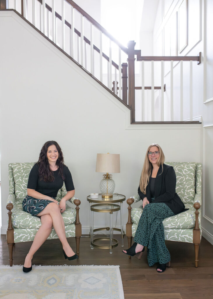Lindsey Putzier, founder and principal designer, and Julianna Watts sitting in the living room on green patterned chairs below the stairs. Chagrin Falls Ohio interior design