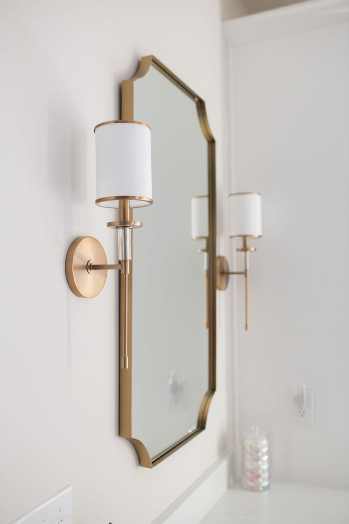 Scalloped mirror in the master bathroom with golden trim and 2 golden sconce lighting fixtures. Lindsey Putzier interior design Peninsula Ohio