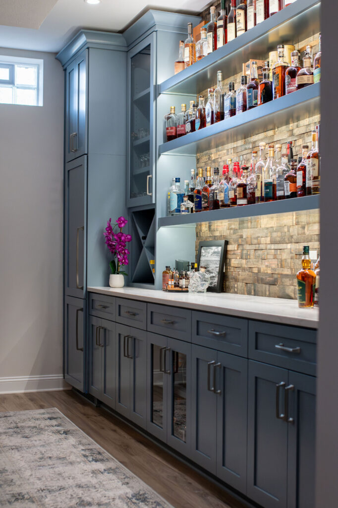 In home bar in the basement with blue custom cabinetry, stone backsplash, under shelving lighting, and marble countertops. Lindsey Putzier interior design 